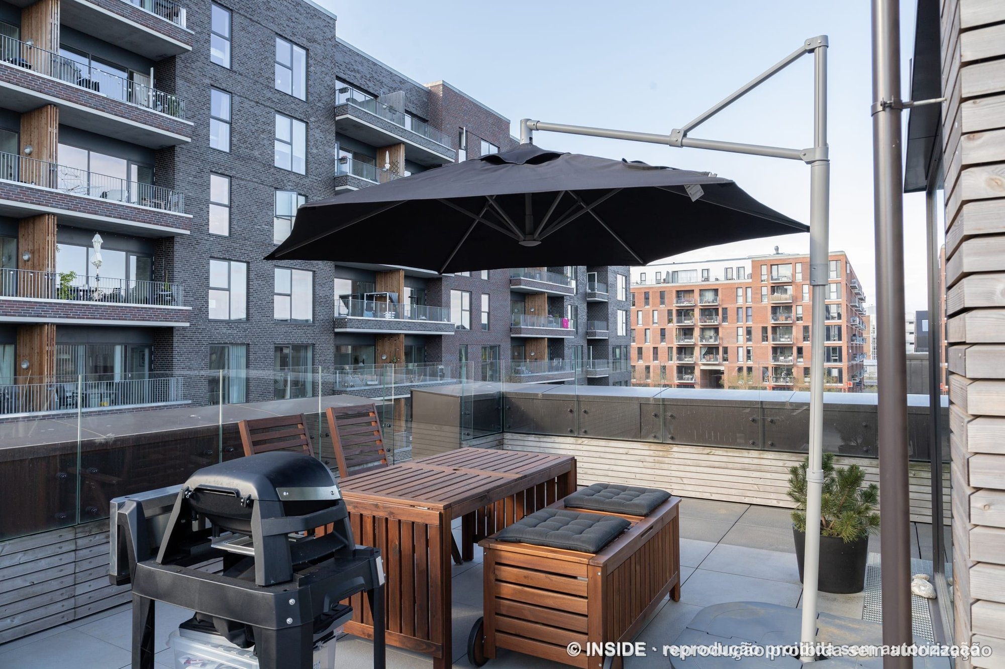 Terraço externo com mesa e bancos de madeira, ombrelone preto, churrasqueira e guarda-corpo de vidro, projeto INSIDE