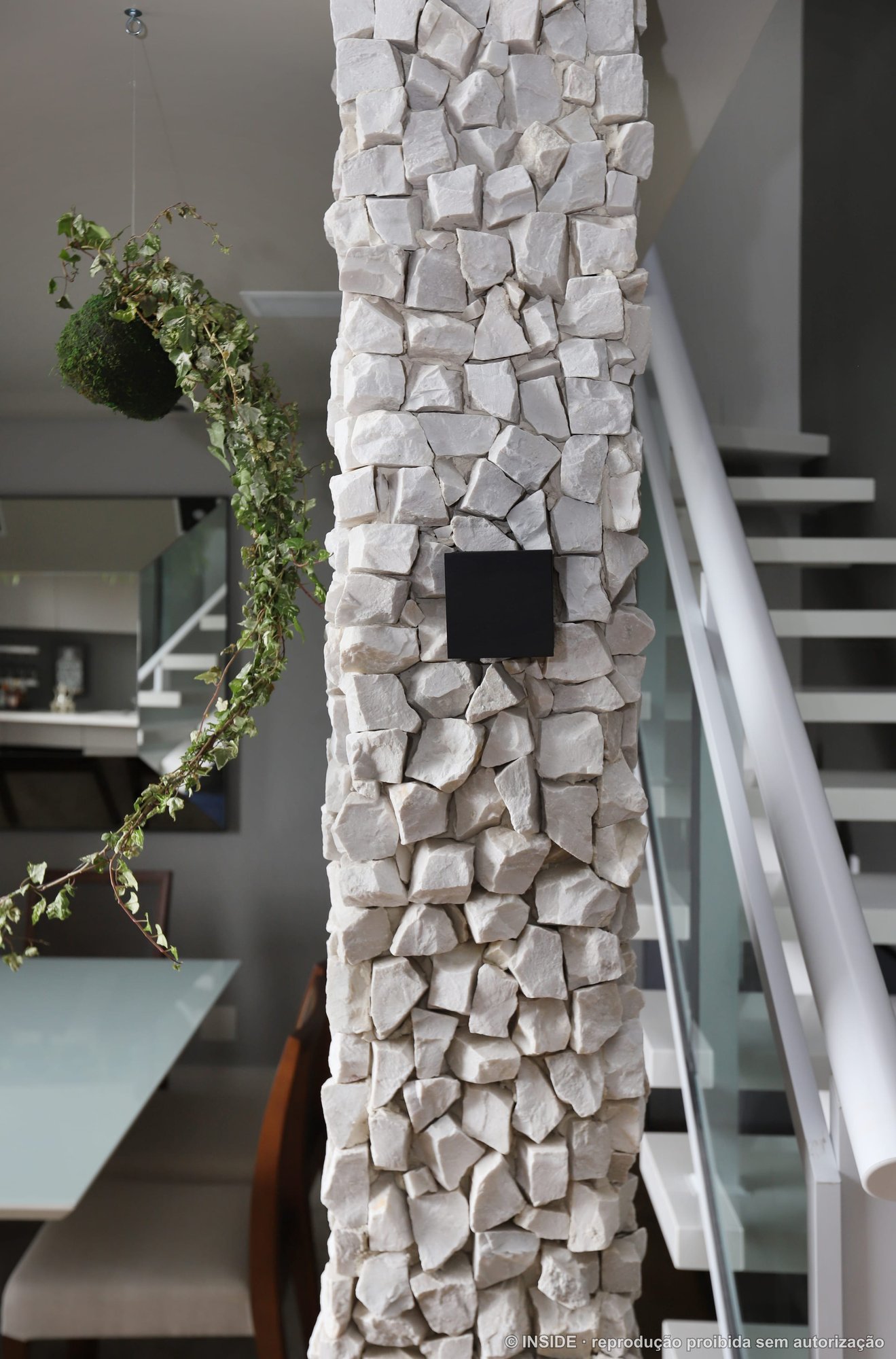 Detalhe da sala com coluna revestida em pedra bruta, escada metálica branca e trepadeira verde descendo pelo pé-direito duplo, projeto INSIDE