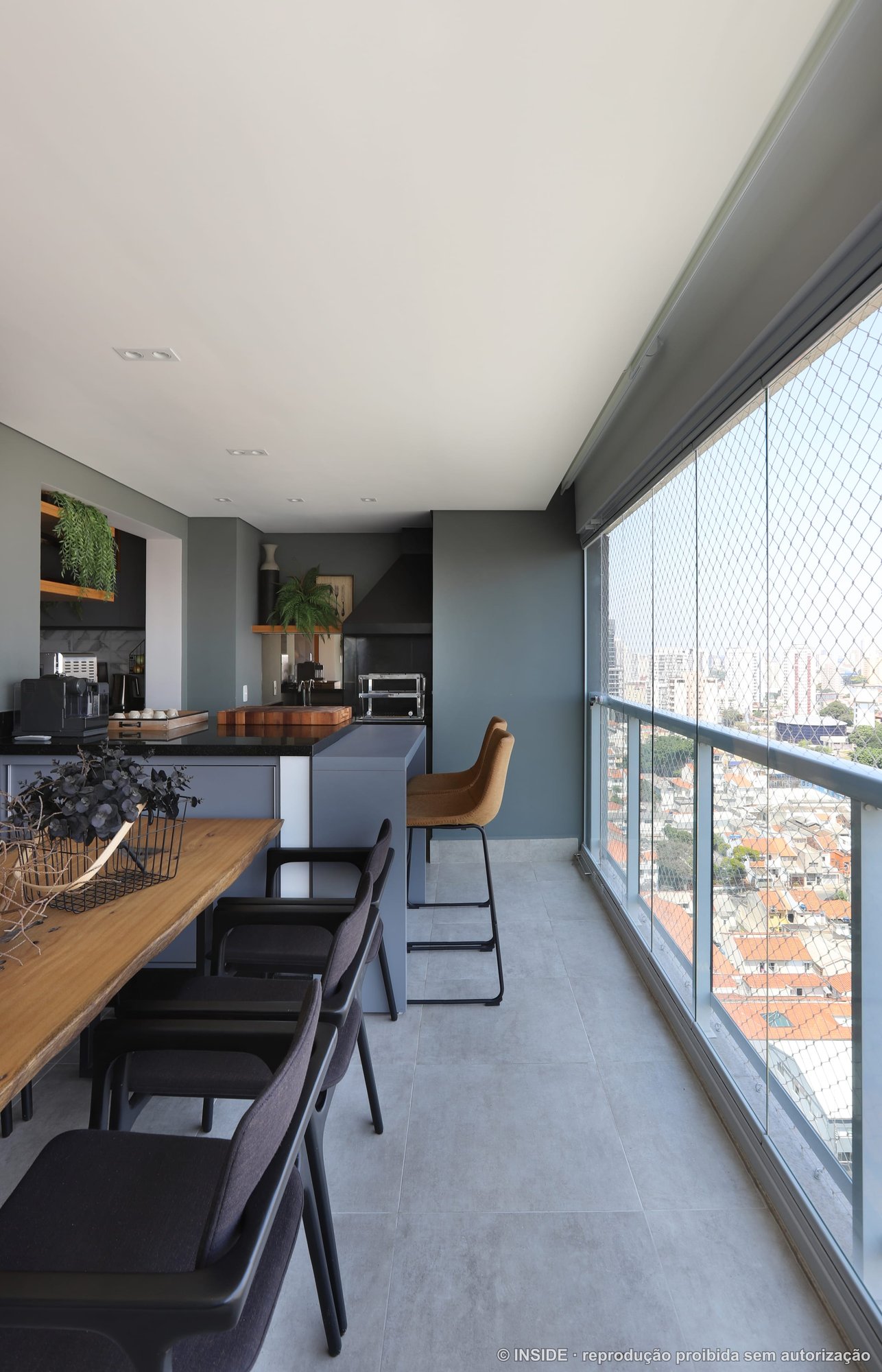 Varanda gourmet com paredes verde escuro, bancada de granito preto, mesa de madeira maciça e vista para a cidade, projeto INSIDE